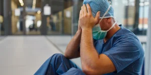 A healthcare worker in scrubs, wearing a surgical mask and hair cover, sits with his head in his hands, appearing distressed or exhausted outside a medical facility.